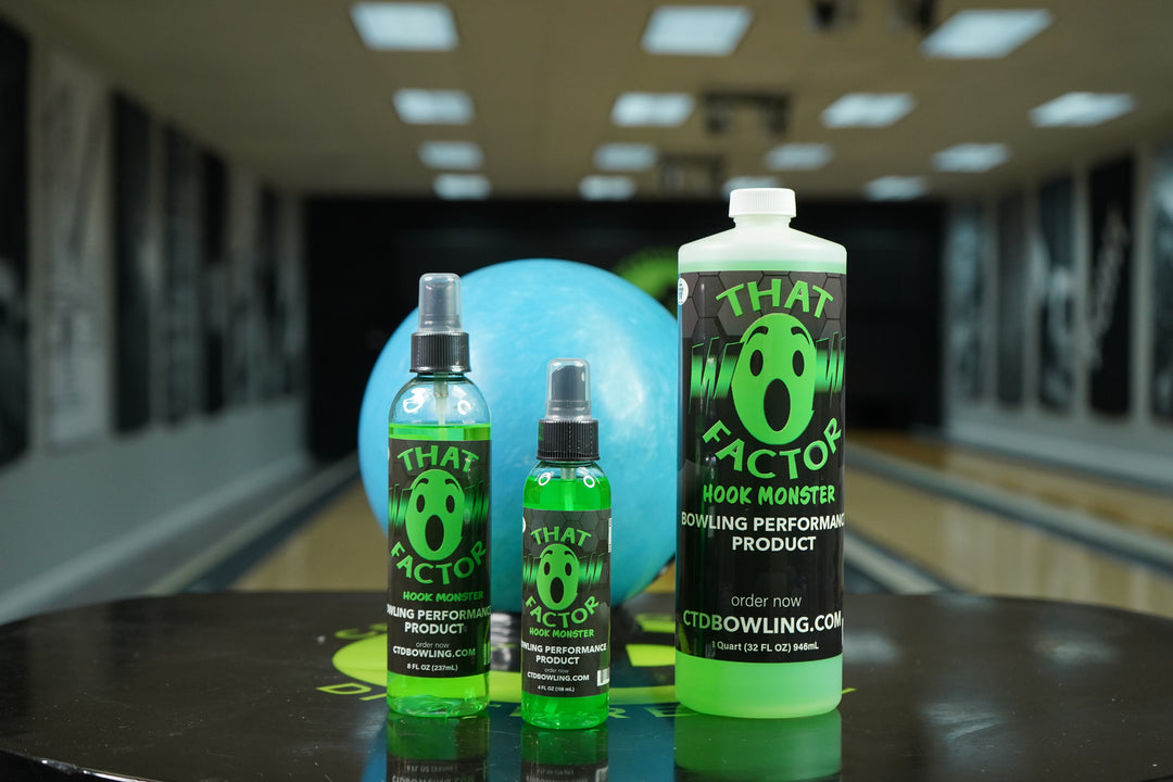 Various sized bottles of That Wow Factor Hook Monster on a table in a bowling alley with a blue bowling ball.