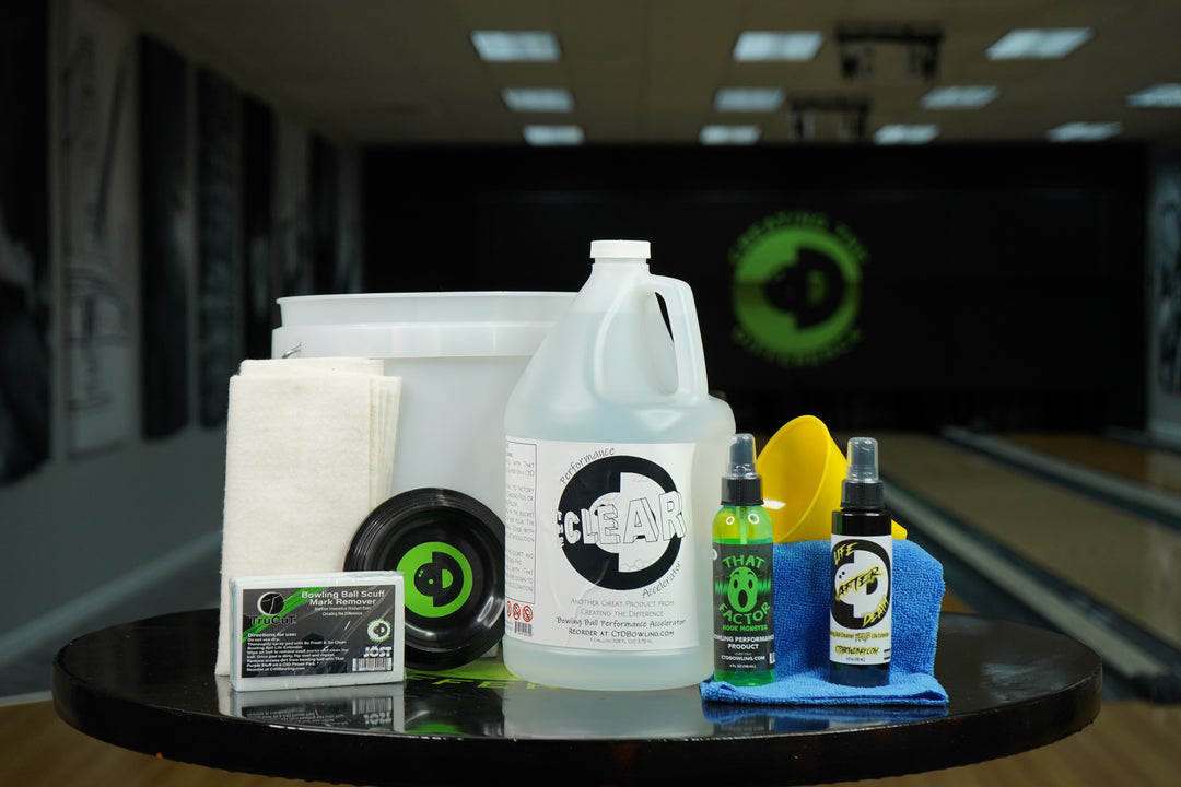 Image of The Clear Ball Restoration Package on a table in a bowling alley. Contains one gallon of The Clear, 4oz each of Life After Death Bowling Ball Cleaner Plus Life Extender and That Wow Factor HM, a CtD Ball Cup, one TruCut Scuff Mark Remover, one microfiber towel, two CtD Absorption Pads, a funnel, and a 2.5 gallon bucket.