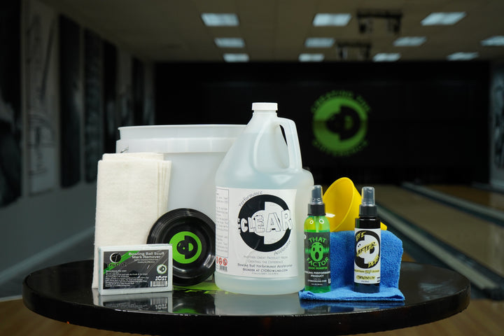 Image of The Clear Ball Restoration Package on a table in a bowling alley. Contains one gallon of The Clear, 4oz each of Life After Death Bowling Ball Cleaner Plus Life Extender and That Wow Factor HM, a CtD Ball Cup, one TruCut Scuff Mark Remover, one microfiber towel, two CtD Absorption Pads, a funnel, and a 2.5 gallon bucket.