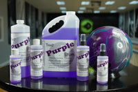 Various size bottles of That Purple Stuff with a bowling ball on a table in a bowling alley